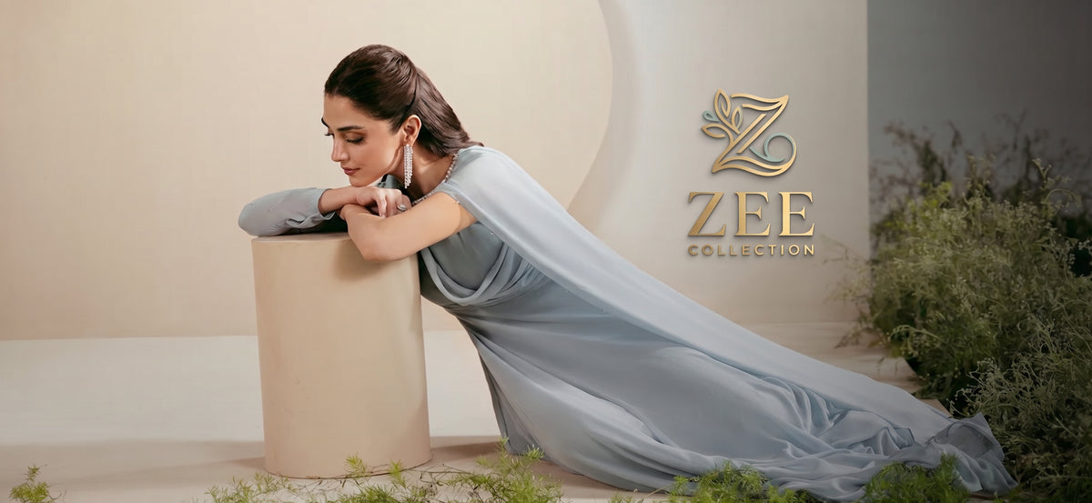 A woman in a flowing light blue dress leans on a beige pedestal in a styled studio setting. The dress has a sheer, draped overlay.