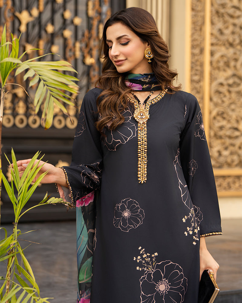 A woman models a black three-piece traditional South Asian salwar suit set with gold floral embroidery, a matching printed dupatta, and a black clutch, posed in front of an ornate golden gate and green plants.